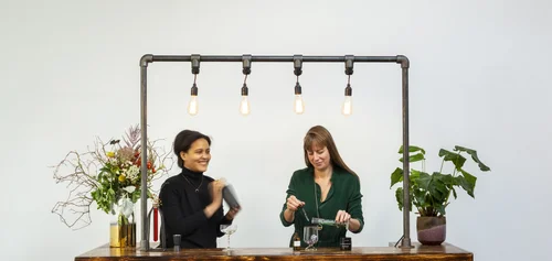 Two guests at the pipe-frame bar, one shaking a cocktail