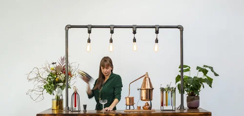 Jemma mixing at the pipe-frame bar with a copper still, flowers and coloured elixir tubes