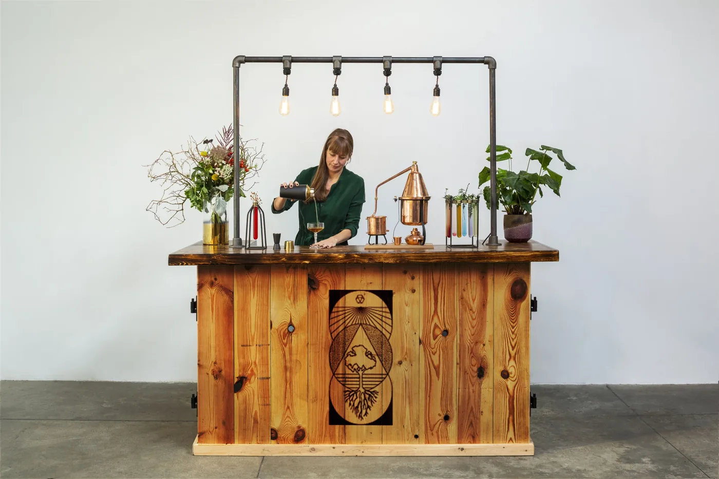 Jemma Foster pouring a cocktail at the reclaimed-wood alchemy bar, with a copper still, Edison bulbs and a sacred-geometry tree-of-life panel