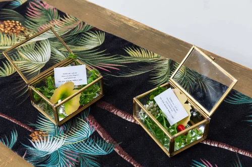 Gold-framed glass boxes of canapés with poem tags on a botanical runner