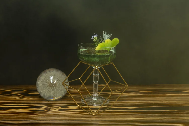 A green herbal cocktail in a coupe on a gold geometric stand beside a dandelion clock
