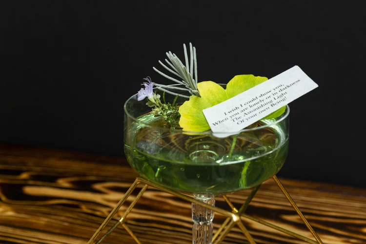 A green cocktail garnished with herbs and a small printed poem